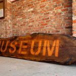 A large wooden sign with the word MUSEUM carved on it rests against a red brick wall near an entrance in Gooding, ID. A framed plaque hangs on the wall above the sign.