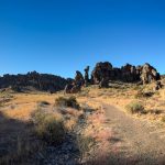A dirt trail winds through a rocky desert landscape near Gooding, ID, with scattered shrubs, rugged cliffs, and rock formations under a clear blue sky, the sun partially visible on the horizon.