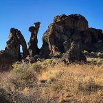 Rock formations rise from dry, grassy terrain under a clear blue sky in Gooding, ID. Sunlight highlights the rugged textures and scattered bushes at the base of the large, weathered rocks.
