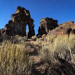 Rock formations with an arch-shaped opening rise against a clear blue sky near Gooding, ID, surrounded by dry shrubs and desert vegetation in the foreground.
