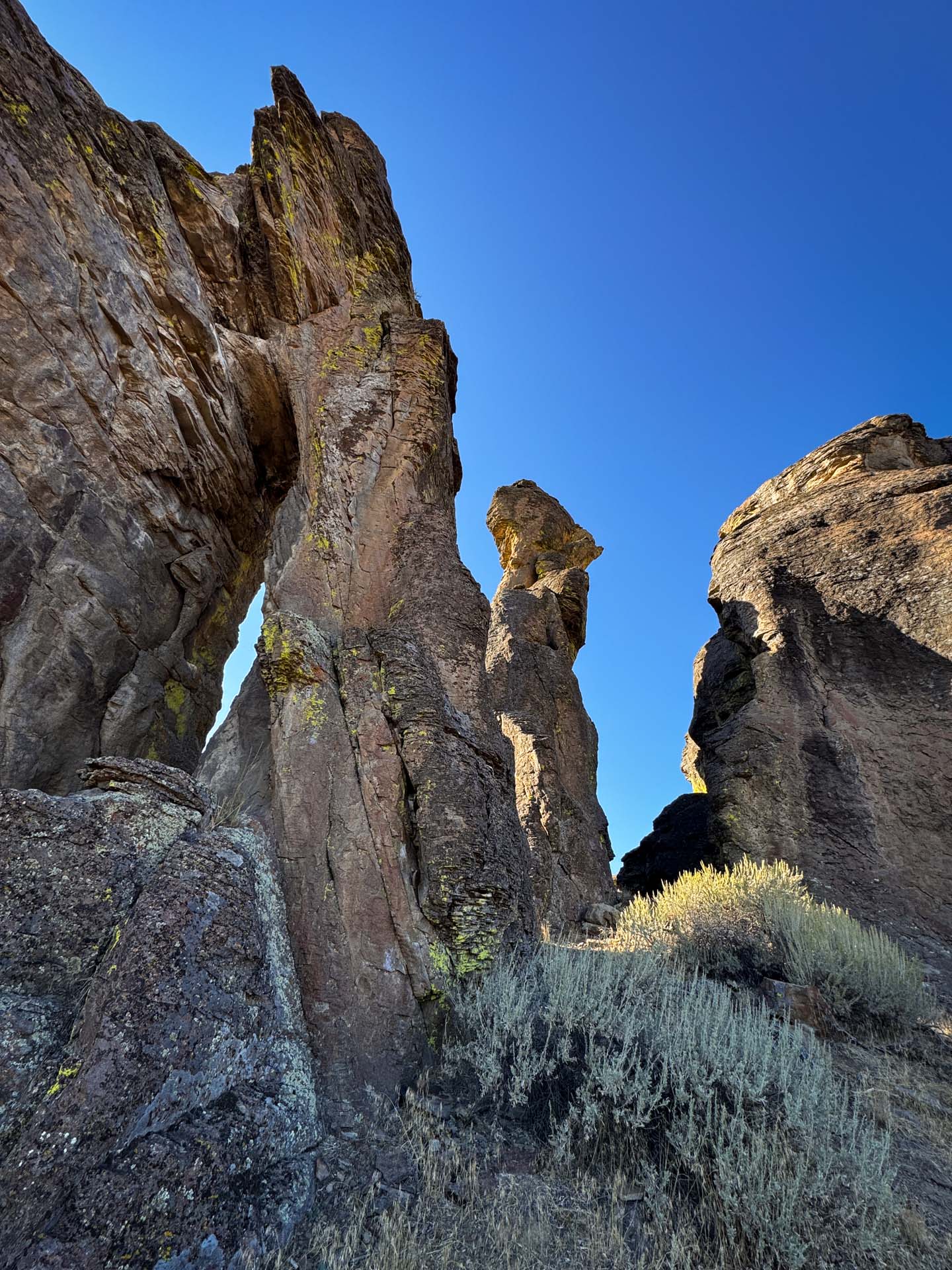 Jagged rock formations rise against a bright blue sky in Gooding, ID, with patches of yellow-green lichen and dry shrubs at the base, creating a dramatic and rugged natural landscape.