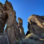 Tall, rugged rock formations rise against a clear blue sky near Gooding, ID, with sparse dry grasses at their base and sunlight casting shadows on the textured surfaces.
