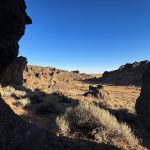 A rocky desert landscape near Gooding, ID, with sparse shrubs and grasses, framed by large dark rocks on both sides, under a clear blue sky.