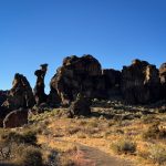 Rock formations rise from a dry, brush-covered desert landscape near Gooding, ID, under a clear blue sky. Shadows cast by the rocks emphasize their rugged shapes and the arid terrain.