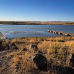 A peaceful lakeside scene near Gooding, ID, with two wooden docks extending into calm water, surrounded by dry grass, rocks, and distant low hills under a clear blue sky.