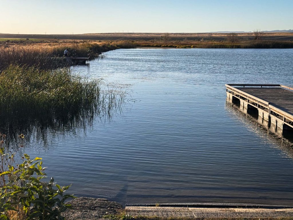 A calm lake in Gooding, ID, bordered by tall grasses and reeds, features two wooden docks stretching into the water from opposite banks beneath a clear sky. The sun casts long shadows across this peaceful scene.