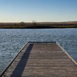 A wooden dock extends over a calm Idaho lake under a clear blue sky, with grassy hills and distant trees visible on the far shore near Gooding.
