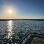 A bright sun shines over a calm, rippling lake near Gooding, ID, with a wooden dock in the foreground. The clear blue sky and sun’s reflection create a shimmering path on the water, while distant land is visible on the horizon.