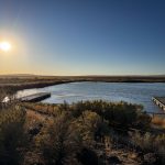 A sunlit lake scene near Gooding, ID, with two wooden docks extending over the water, surrounded by bushes and grasses, under a clear blue sky during late afternoon or early evening.