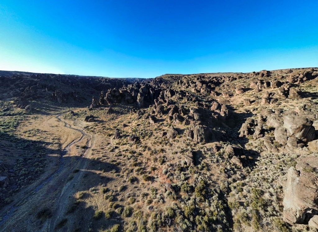 Wide landscape of a rocky desert near Gooding, ID, with scattered shrubs, rugged terrain, and a dirt path winding through the area under a clear blue sky. Shadows from rock formations stretch across the ground.