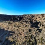 Wide landscape of a rocky desert near Gooding, ID, with scattered shrubs, rugged terrain, and a dirt path winding through the area under a clear blue sky. Shadows from rock formations stretch across the ground.