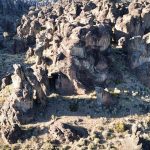 Aerial view of a rugged desert landscape in Gooding, ID, with tall, jagged rock formations, sparse vegetation, and a dirt path winding through the area under bright sunlight.