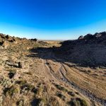 A wide, rocky desert landscape near Gooding, ID, with sparse vegetation, rugged hills, and a dirt path winding through the terrain under a clear blue sky.