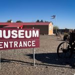 A red and white sign reading MUSEUM ENTRANCE stands on a gravel area in Gooding, ID, with an old wagon and a building with a red roof in the background under a clear blue sky.