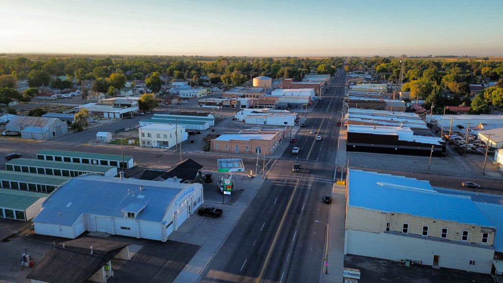 Aerial view of Gooding, ID with low-rise buildings, tree-lined streets, and a few cars on the main road at sunset. The surrounding area is flat and green.