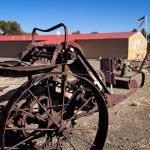 An old, rusty piece of farm equipment with large metal wheels sits on gravel in front of a tan building with a red roof and murals in Gooding, ID, under a clear blue sky.