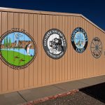 A tan metal building wall in Gooding, ID, displays five colorful circular emblems representing the City of Hagerman, Gooding Idaho, Gooding County Idaho, and local heritage symbols under a clear blue sky.