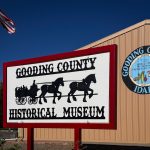 A sign for the Gooding County Historical Museum in Gooding, ID features a horse-drawn wagon graphic. Behind it is a building with a round seal that says “Gooding County Idaho.” An American flag is visible at the left.