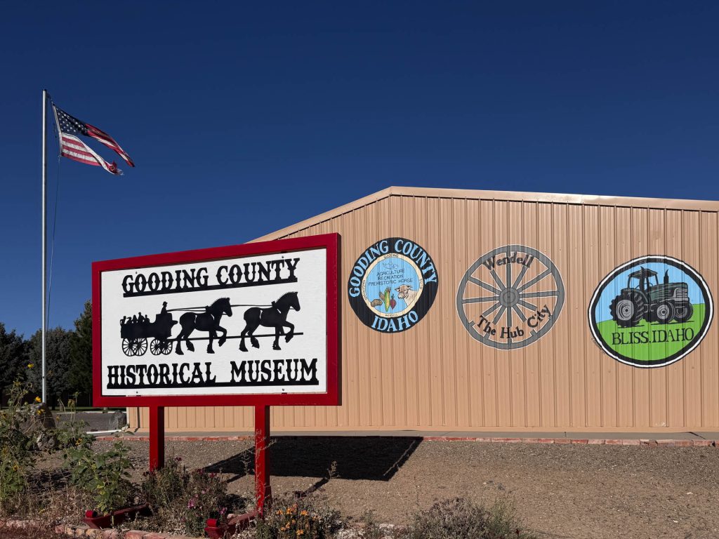 A beige building in Gooding, ID with three circular murals, a large sign reading Gooding County Historical Museum, and an American flag flying on a pole under a clear blue sky.