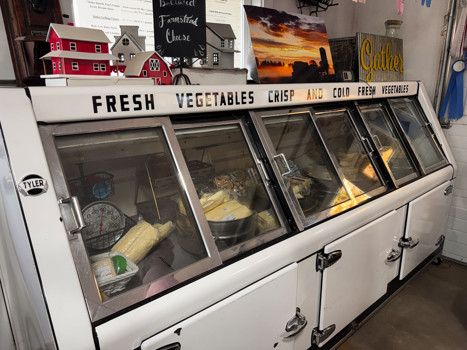 A vintage refrigerated display case labeled Fresh Vegetables holds ears of corn and other produce in a rustic Gooding, ID indoor setting. Decorative signs, a scale, and model houses sit atop the case, adding to the charming atmosphere.